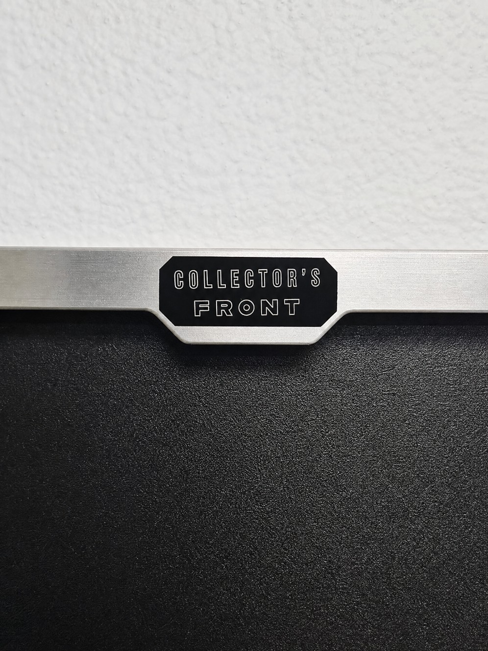 Close-up of Collectors Front engraved aluminum badge on display frame showing brushed aluminum finish and black Sintra board base