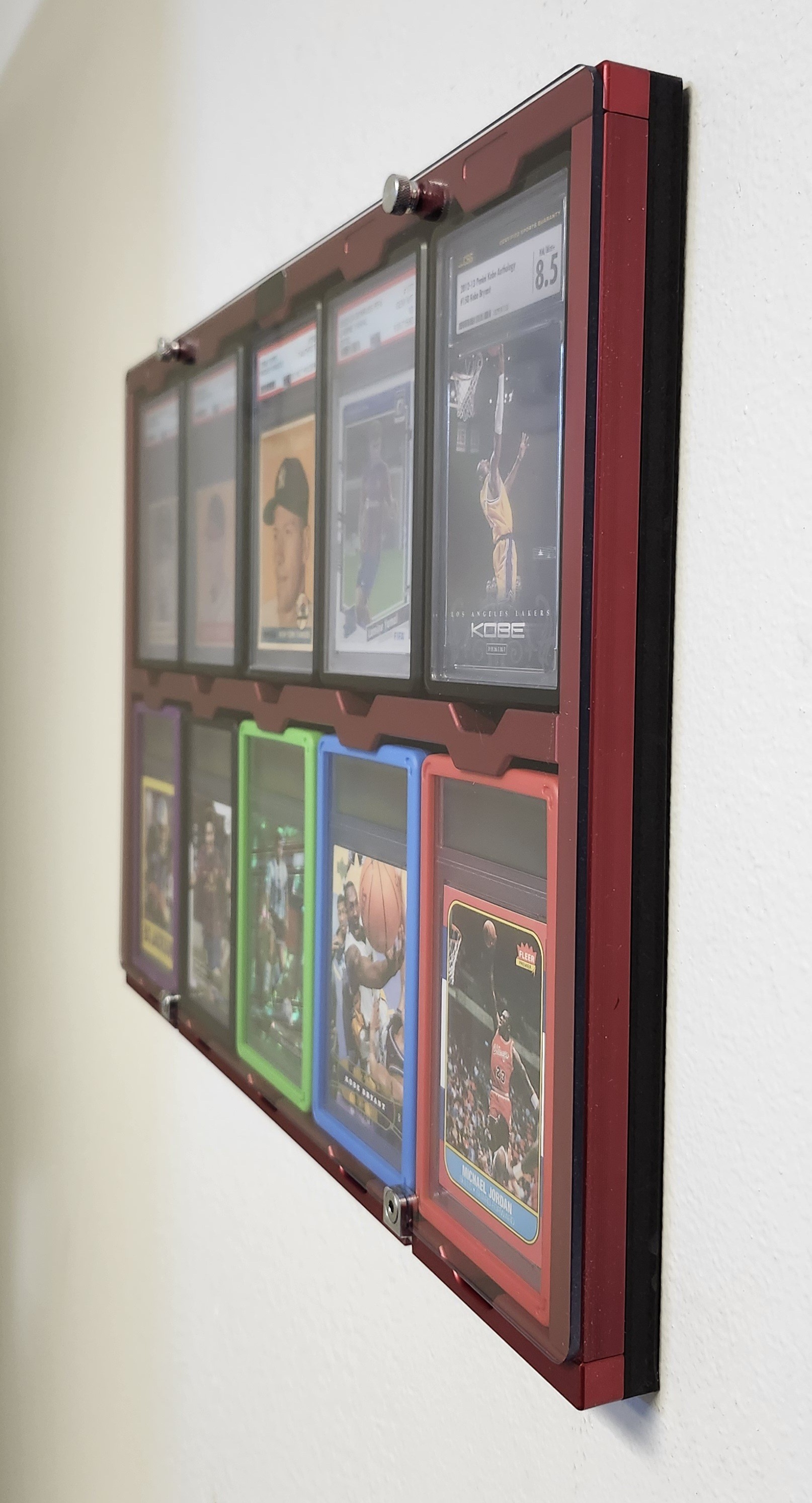 Red anodized Protected Display side angle view with colorful slab bumpers showing UV polycarbonate window and graded cards including Kobe Bryant and Michael Jordan